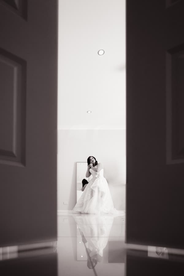 The bride Holly adjusts her earring while standing in front of a mirror at Kwila Lodge, visible through a partially open door.