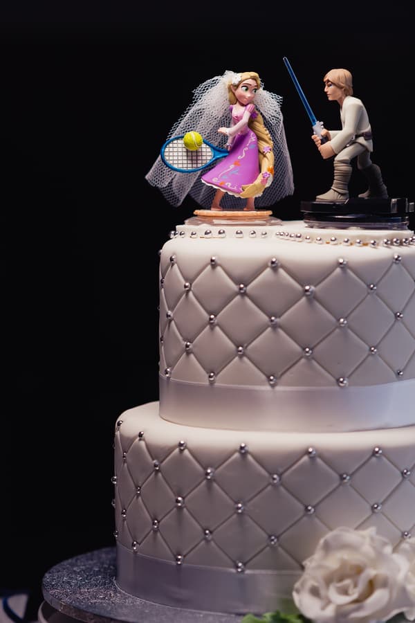 Wedding cake at TOOWONG ROWING CLUB — The Malouf Room with a quilted white fondant design and silver bead accents, topped with figurines of a bride holding a tennis racket and a groom holding a blue lightsaber.