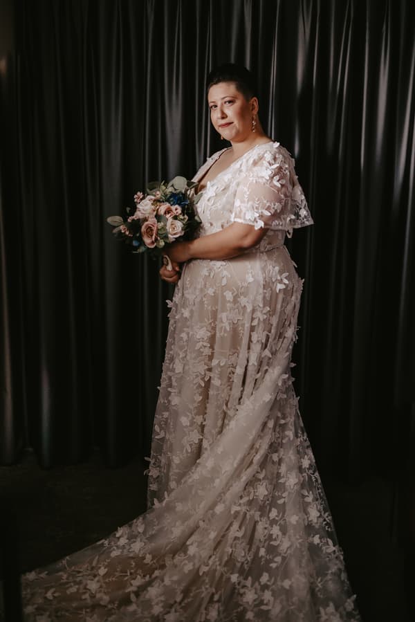 Bride in a white floral lace wedding gown holding a bouquet of flowers, standing in front of dark curtains.