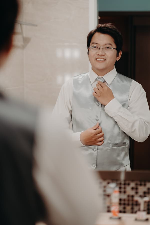 The groom Timothy adjusts his tie and buttons his silver vest while looking into a mirror at Royal on the Park.