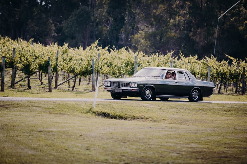 The bride and groom are seated inside a vintage car parked on a grassy area near a vineyard at Ocean View Estates.