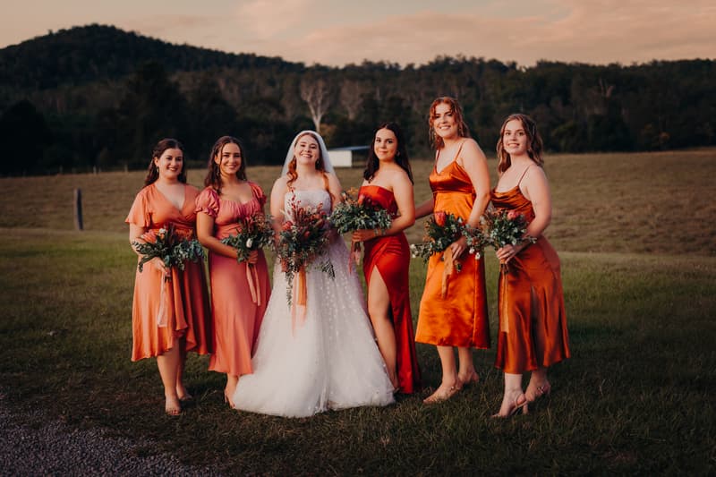 The bride Lilly stands with five bridesmaids in various shades of orange and red dresses holding bouquets at Yabbaloumba Retreat — By The River with a grassy field and hills in the background.