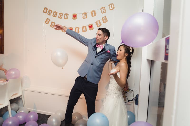 Jason and Wing pose playfully on the reception stage at Bilinga Beach Weddings — Bilinga SLSC, surrounded by pastel balloons and a banner displaying their names and wedding date.