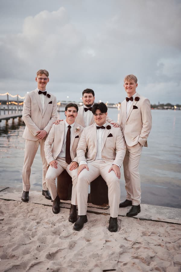 Joel and four groomsmen pose together on the sandy shore at Sandstone Point Hotel, with a pier and water in the background during the couple portraits stage.