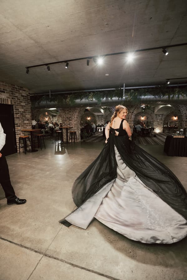 The bride Jayme twirls in her wedding dress with a long black overlay train at the reception stage inside Sandstone Point Hotel — The Cellar, with guests seated at tables in the background.