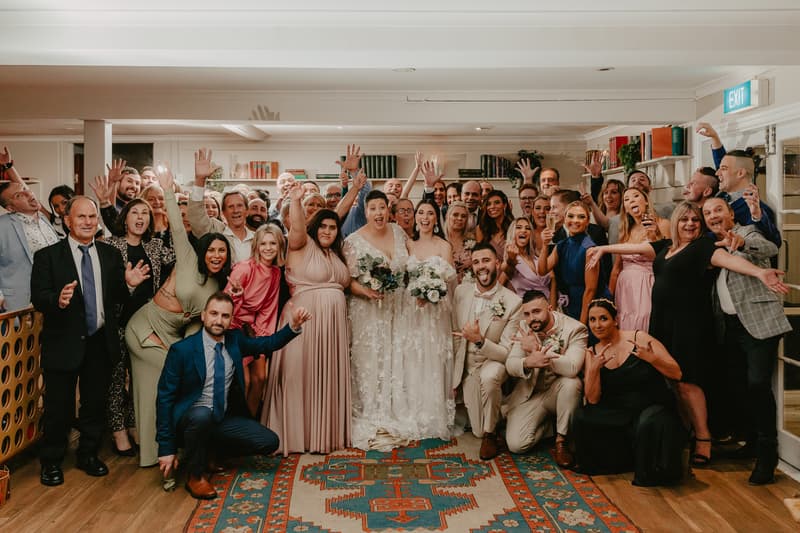 Large group photo with two brides in white wedding dresses holding bouquets, surrounded by guests and wedding party members indoors.