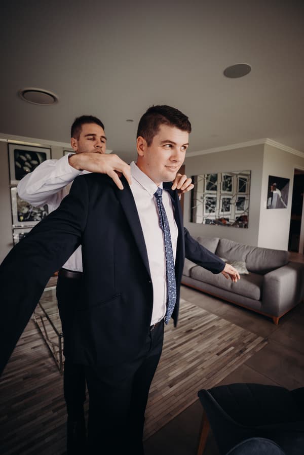 The groom Brent is being helped to put on his suit jacket by a groomsman in a living room setting at Preston Peak Winery Functions Centre.