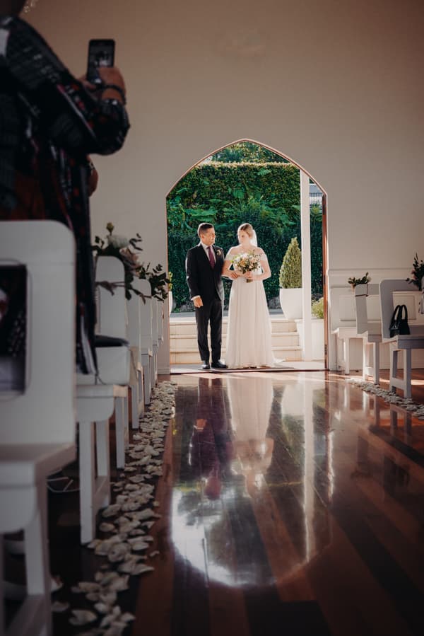 Courtney the bride and Liam the groom stand together at the entrance of Tiffany's Maleny chapel, with Courtney holding a bouquet and guests seated along the aisle decorated with flower petals.