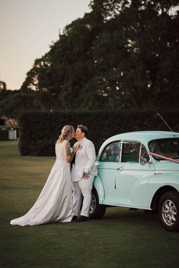 Mandy and Selina share a kiss beside a vintage light blue car on the lawn at Sandstone Point Hotel during their couple portraits session.