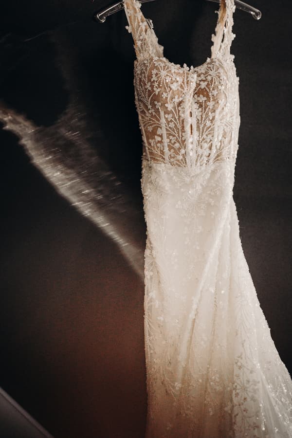 The bride's wedding dress with intricate floral lace and bead detailing hanging on a hanger against a dark background.