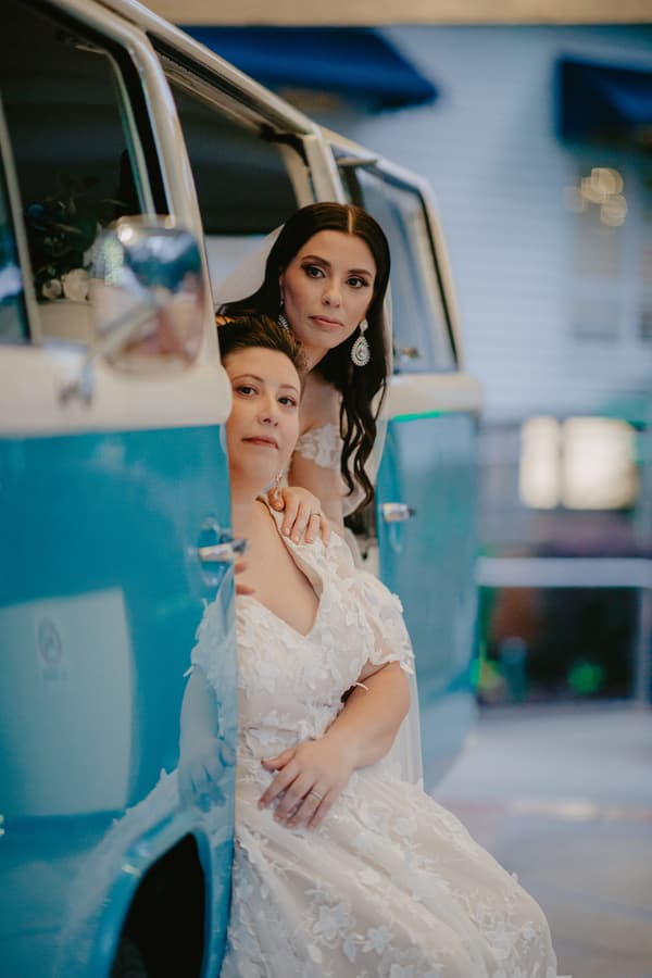 Two brides in white lace wedding dresses are seated in and leaning out of the open door of a blue vintage van.