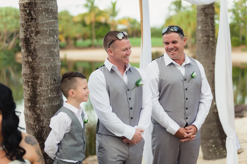 Two groomsmen and a ring bearer stand smiling near palm trees at the ceremony stage at Novotel Sunshine Coast Resort.