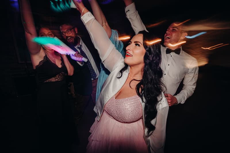 The bride Mindy in a pink dress and white jacket dances with the groom David in a white shirt and black bow tie, along with other guests, at the reception stage in Sandstone Point Hotel — Cellar.