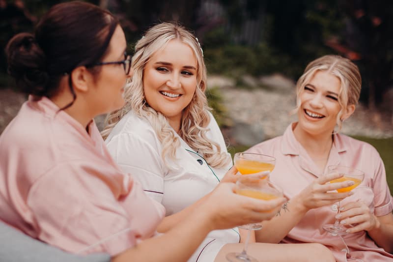 The bride Chloe in white pajamas and two bridesmaids in pink pajamas sit outdoors holding glasses of orange juice, smiling and chatting.