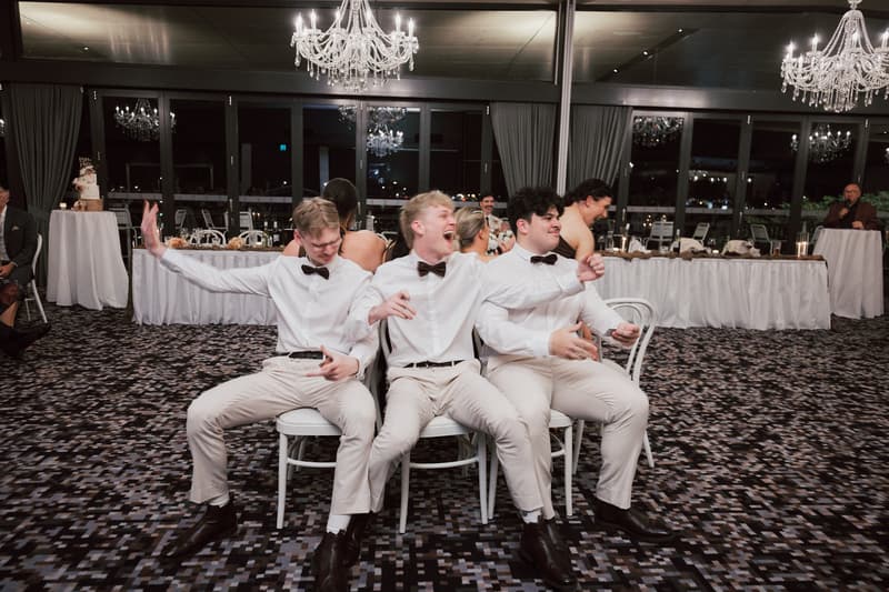Three groomsmen in white shirts and bow ties sit back-to-back on chairs playing a game at the reception in The Pumicestone Room, Sandstone Point Hotel, with guests and a speaker visible in the background.