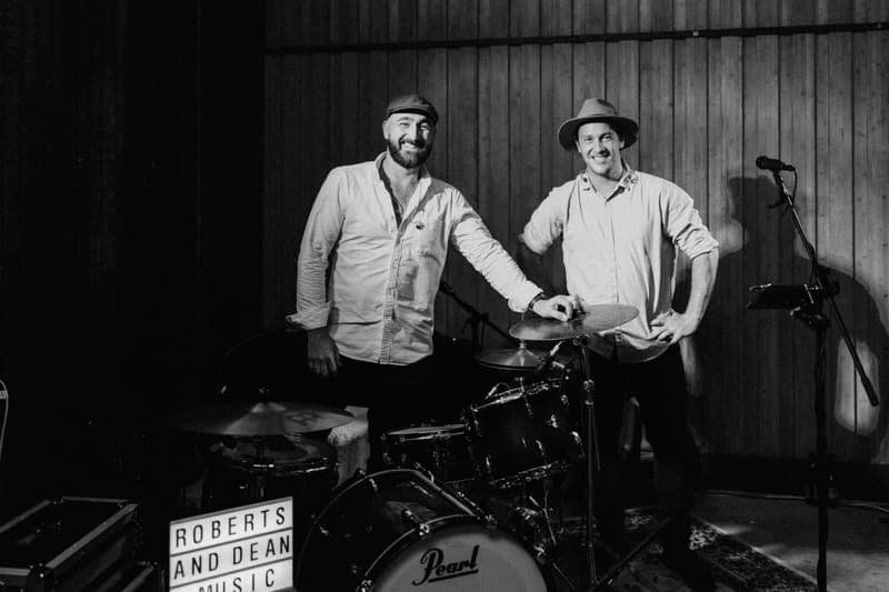 Two men stand behind a drum set and microphone stand at Yabbaloumba Retreat — The Shed, with a lightbox sign reading 'ROBERTS AND DEAN MUSIC'.