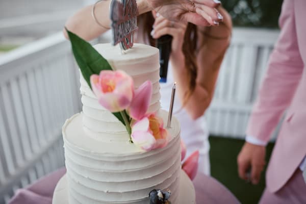 The bride and groom at White Horse Ranch prepare to cut a white tiered wedding cake decorated with pink flowers and a lit candle on top.