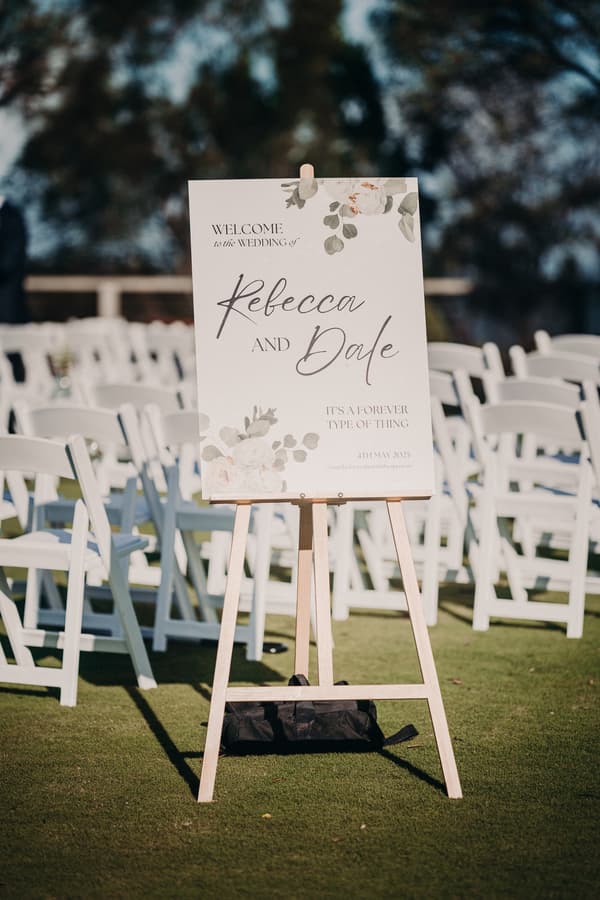 Welcome sign for the wedding of Rebecca and Dale displayed on a wooden easel at Sandstone Point Hotel with rows of white folding chairs arranged on grass in the background.
