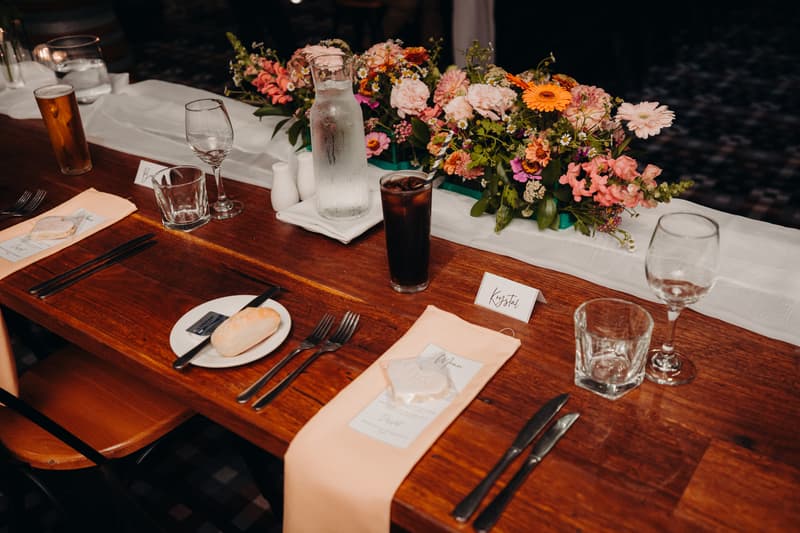 Reception table setting at Sandstone Point Hotel — Cellar with floral centerpiece, place cards including one for the bride Krystal, glassware, cutlery, napkins, and beverages.