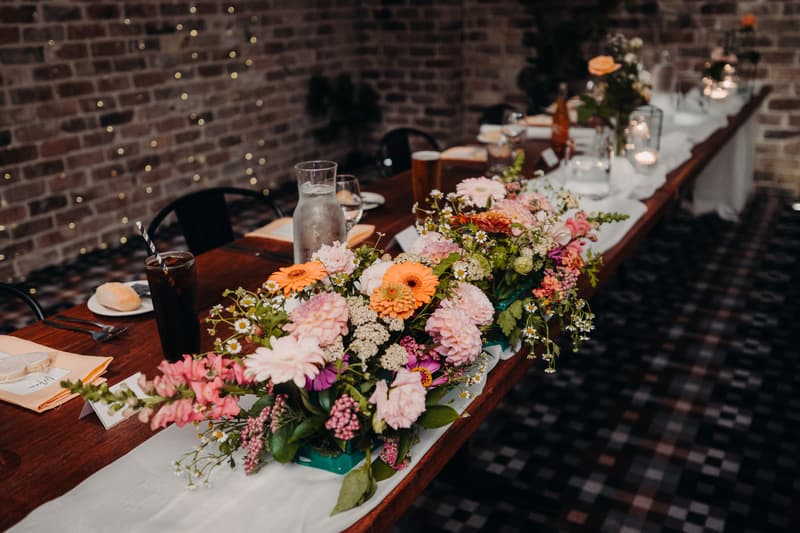 Long wooden table at Sandstone Point Hotel — Cellar reception stage decorated with floral centerpieces, glassware, plates, and drinks.