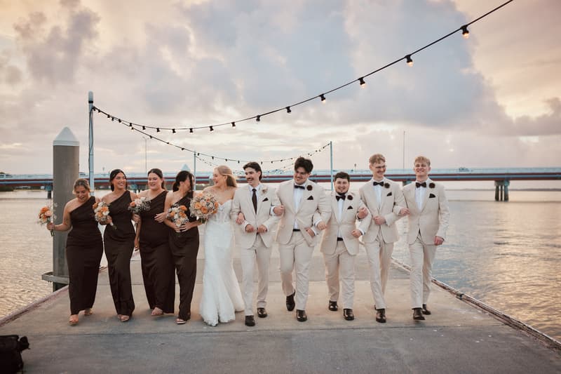 The bride Morgan and groom Joel walk arm-in-arm with their bridal party on a pier at Sandstone Point Hotel, with bridesmaids in dark dresses holding bouquets and groomsmen in light suits with bow ties, under string lights at sunset.