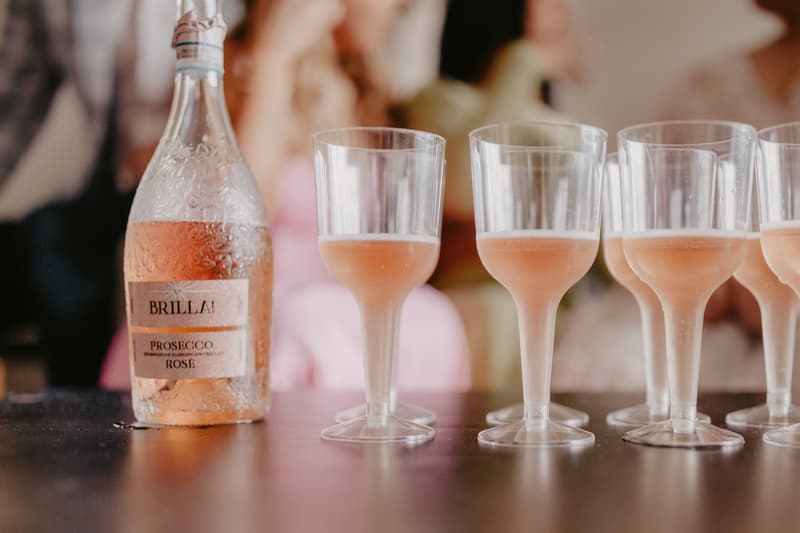 A bottle of Brilla! Prosecco Rosé and six plastic glasses partially filled with rosé prosecco on a dark surface.