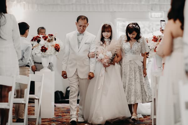 The bride walks down the aisle at Royal on the Park, accompanied by a man in a white suit and a woman in a floral dress, with guests seated on either side and floral arrangements lining the aisle.