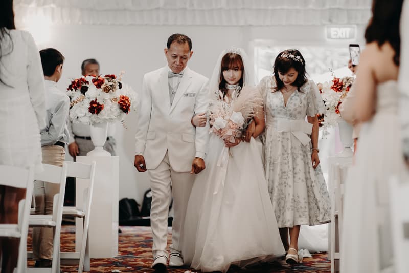 The bride walks down the aisle at Royal on the Park, accompanied by a man in a white suit and a woman in a floral dress, with guests seated on either side and floral arrangements lining the aisle.