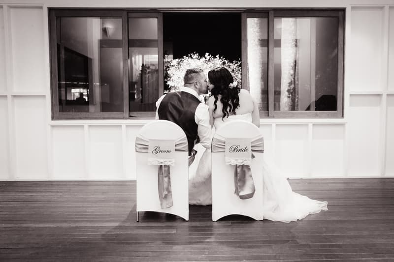 The bride and groom sit side by side on chairs labeled 'Bride' and 'Groom' at Novotel Sunshine Coast Resort, sharing a kiss with their backs to the camera in front of a large window.
