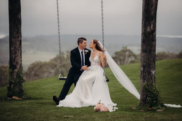 Bride Chelsea and groom Brent sit together on a swing outdoors at Preston Peak Winery Functions Centre with a scenic landscape in the background. Chelsea wears a white wedding dress with a long veil and holds the swing chains, while Brent wears a dark suit with a boutonniere. A bouquet of flowers lies on the grass in front of them.