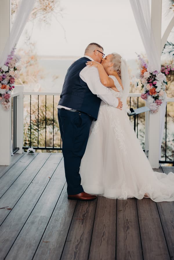 Bride Chantelle and groom Peter share a kiss during their ceremony at Sandstone Point Hotel — Pavilion, framed by floral arrangements and white drapery.