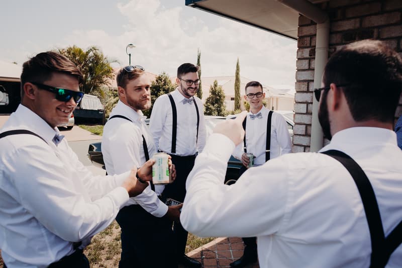 Five groomsmen dressed in white shirts with black suspenders and bow ties stand outside near a brick building, holding canned drinks and engaging in conversation.