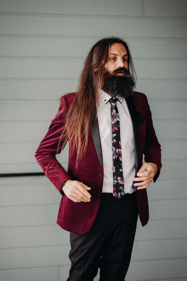 The groom stands alone wearing a burgundy velvet jacket, white shirt, and floral tie against a light gray paneled wall at Sandstone Point Hotel.