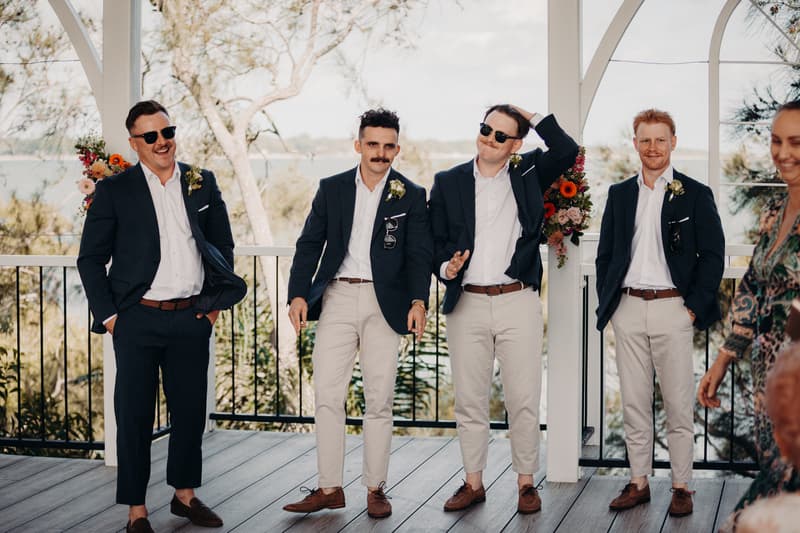 Four groomsmen stand on the ceremony stage at Sandstone Point Hotel — Pavilion, dressed in navy blazers, white shirts, and light-colored trousers with boutonnieres, two wearing sunglasses.