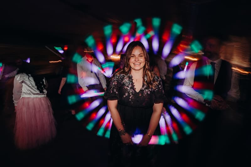 A female guest in a black floral dress stands smiling on the reception stage at Sandstone Point Hotel — Cellar, with colorful circular light effects behind her and other guests dancing in the background.