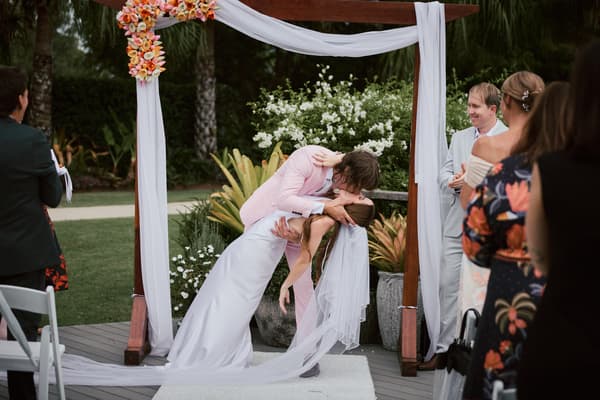 Olivia and Jake share a kiss under a floral arch at the ceremony stage at Eatons Hill Hotel — Lakeside, surrounded by guests watching and smiling.