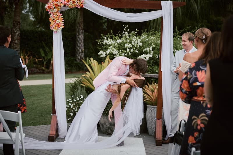 Olivia and Jake share a kiss under a floral arch at the ceremony stage at Eatons Hill Hotel — Lakeside, surrounded by guests watching and smiling.