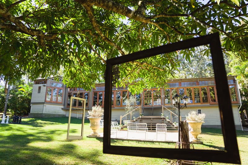 Exterior view of Kwila Lodge showing a large veranda with stairs, white outdoor chairs, large planters, and a black lamp post, framed by two empty picture frames hanging from a tree branch.