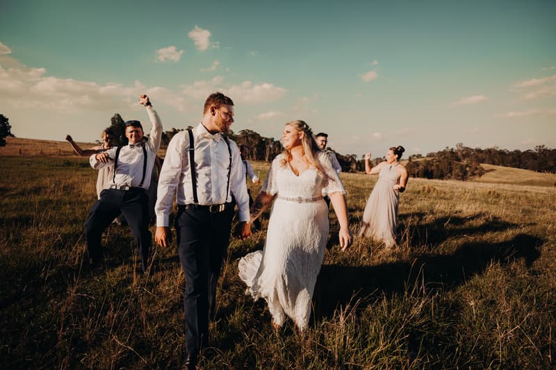 Bride Annie and groom Trent walk hand in hand through a grassy field at Ocean View Estates, followed by bridesmaids and groomsmen in formal attire.