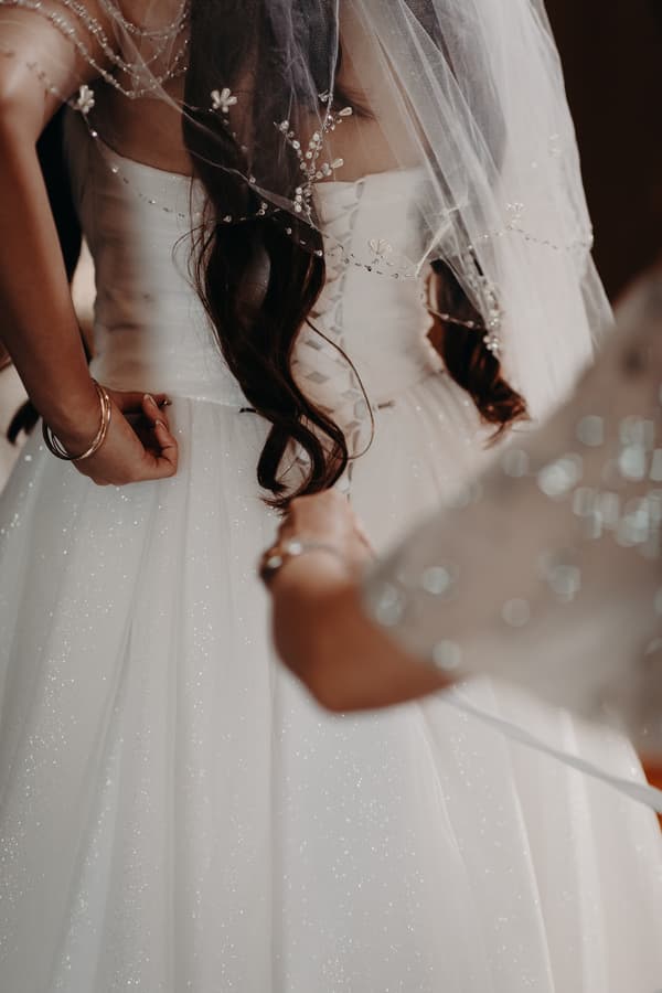 The bride at Royal on the Park is having the back of her wedding dress laced up by another person, showing the detailed veil and sparkling fabric of the gown.