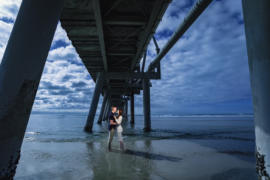 The couple stands under a pier at The Spit, Gold Coast, embracing with a dramatic cloudy sky above.