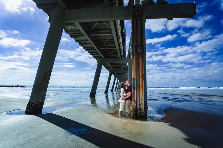 The couple leans against a pier column at The Spit, Gold Coast, with waves in the background and bright sunlight.