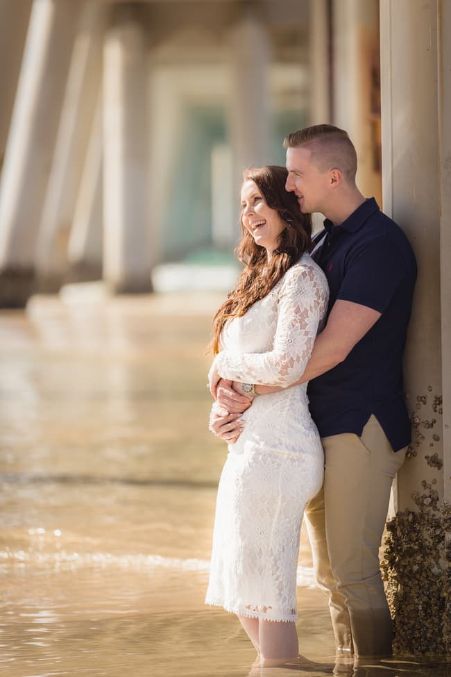 The couple stands close, smiling and embracing by a pier column at The Spit, Gold Coast, in soft light.