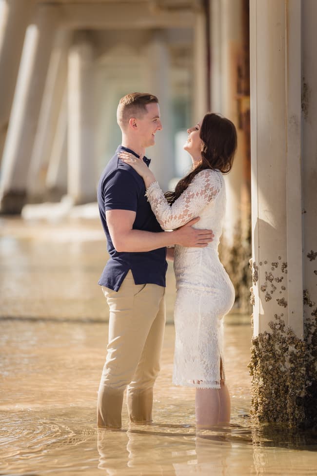 The couple gazes at each other, standing in shallow water by a pier at The Spit, Gold Coast, under soft lighting.