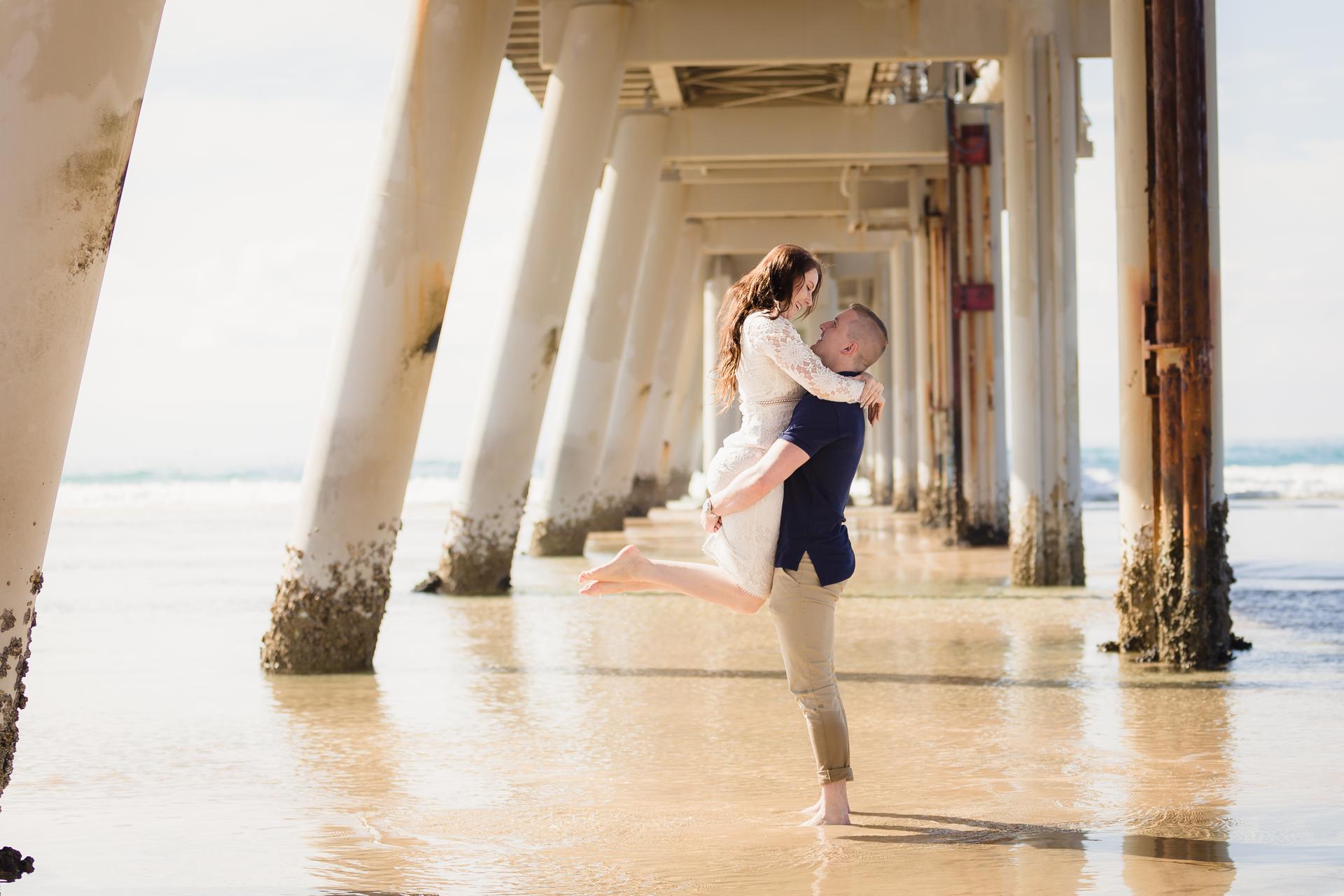 The couple shares a playful lift under the pier at The Spit, Gold Coast, with sunlight streaming through.