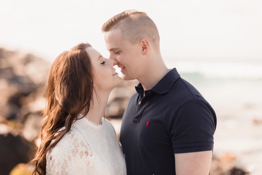 Close-up of the couple almost kissing, standing on a beach with blurred waves in the background.