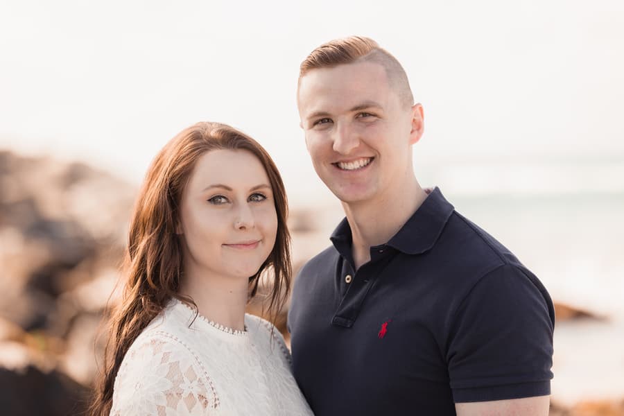The couple smiles warmly at the camera, standing on a beach with rocks and ocean waves in the background.