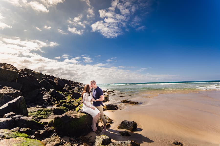 The couple sits on rocks by the beach at The Spit, Gold Coast, with a bright blue sky and ocean in the background.
