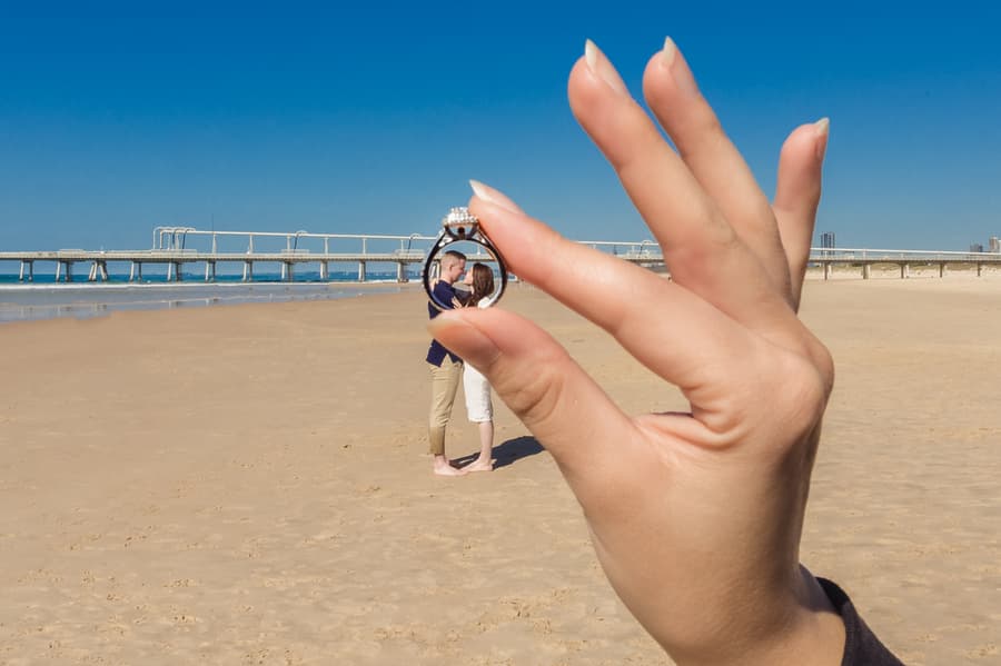 A hand holds an engagement ring in focus, with the couple blurred in the background on a sandy beach.