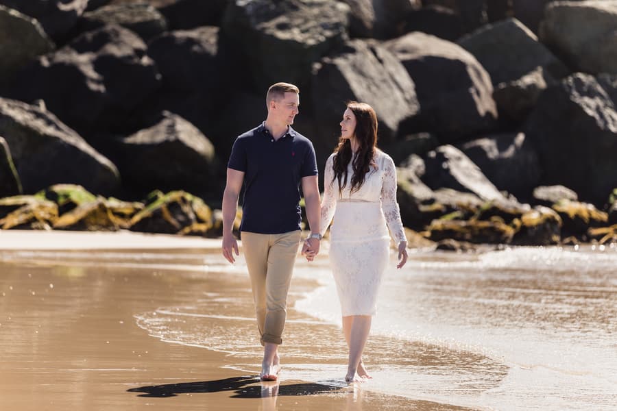 The couple walks hand in hand along the shoreline at The Spit, Gold Coast, with rocky cliffs in the background.
